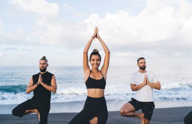 Sahilde bütünsel iyileşme sırasında kameraya gülümseyen günlük eşofmanlı neşeli kadın yogi portresi, kıyı şeridinde erkeklerle yoga yaparken ince vücutlu poz veren mutlu kadın.