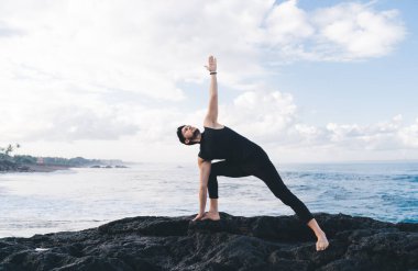 Sportif giyinen sakin bir erkek yoga pozisyonunda duruyor ve sabah bilinç yoğunluğu sırasında deniz kıyısında meditasyon yapıyor. Kafkasyalı bir adam aydınlanma uyumu için hatha çalışmasının keyfini çıkarıyor.