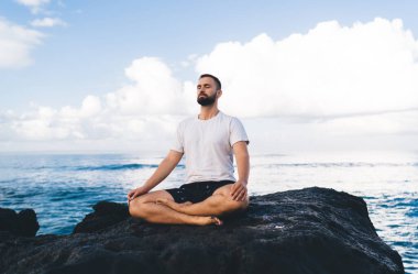 Kafkas erkek yogi rahat giyinmiş, lotus şeklinde meditasyon yapıyor, deniz kıyısındaki kayalarda rahatlama ve konsantrasyon zamanının tadını çıkarıyor, bütünsel iyileşme kavramı ve zihinsel sağlıklı yaşam tarzı.