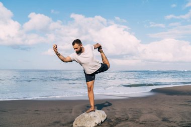 Esnek bir adam dengede duruyor Asana 'yı sabah mavi gökyüzü ile kıyı şeridinde yükselen bacak nefesi, bir bacağının üzerinde duran konsantre bir adam deniz kıyısı egzersizleri sırasında egzersiz yapıyor.