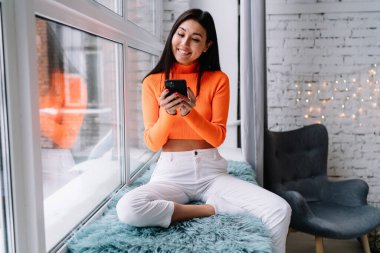 Genç gülümseyen Asyalı kadın pencere pervazında kürklü halıda oturuyor ve akıllı telefondan bir şeyler izliyor. Ev hayatı kavramı. Bin yıllık neslin güzel esmer kızı. Modern daire