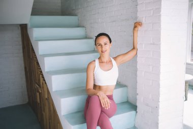 Genç beyaz kız merdivenlerde oturuyor ve geniş evdeki kameraya bakıyor. Modern kadın yaşam tarzı kavramı. Gülümseyen esmer kadın spor giysisi ve yalınayak. Güneşli gündüz