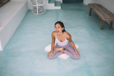 Balili kız geniş bir evde yoga yapıyor. Sağlıklı yaşam tarzı kavramı. Ağzı açık, spor giyimli ve çıplak ayaklı genç bir kadın yastığa oturmuş kameraya bakıyor.