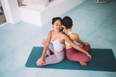 Kız arkadaşlar geniş evde yoga yaparken Twist pozu veriyorlar. Sağlıklı yaşam tarzı kavramı. Arkadaşlık fikri. Spor giyimli ve çıplak ayaklı kadınların spor minderinde sırt sırta oturuşunun en iyi görüntüsü.