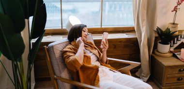 Asyalı kız evdeki pencerenin yanındaki koltukta oturup dinlenirken akıllı telefondan bir şey izliyor. Ev hayatı kavramı. Gündelik kıyafetler giyen genç bir kadın. Modern apartman içi