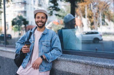 Turistik şehirde kahve molası sırasında kameraya gülümseyen modaya uygun kot giymiş neşeli hippi bir adamın yarım boy portresi.