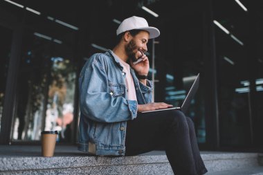 Neşeli hipster adam dizüstü bilgisayarda kurgulanmış içerik vlog 'unu izlerken meslektaşını fikirleri tartışmaktan vazgeçirmek için arıyor, mutlu milenyum serbest çalışanı telefon için cep telefonu ve ağ kurmak için ağ defteri kullanıyor.