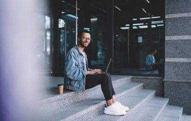 Optik gözlüklü neşeli hipster adamın portresi kamera karşısında gülümseyen göz düzeltmesi sağlıyor netbook üzerinde uzaktan çalışan mutlu erkek blogcu şehir merdivenlerinde dizüstü bilgisayarla poz veriyor.