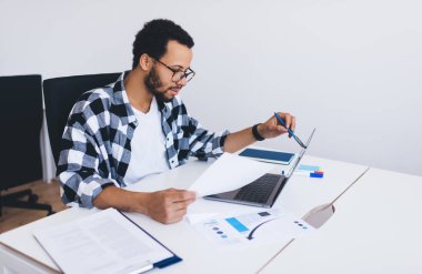 Ofis içi mali analiz sırasında kağıt ve web raporlarını kontrol eden optik gözlüklü yetenekli erkek çalışan, çalışma sırasında dizüstü bilgisayar kullanan gözlüklü bir milenyum adamı.