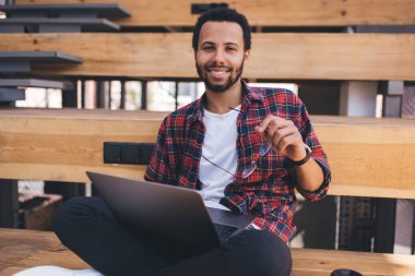 Netbook 'ta uzaktan çalışan neşeli genç bir programcının portresi. Serbest yaşam tarzının tadını çıkaran mutlu hipster adam dizüstü bilgisayardaki yazılımı güncellerken kameraya gülümsüyor ve 4G kablosuz kullanıyor.