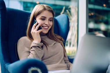 Telefondan konuşan, gülümseyen, rahat mavi koltukta oturan ve geniş pencereli parlak salonda dizüstü bilgisayarla çalışan güzel bir kadın.