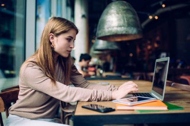 Sınavlara hazırlanırken pencerenin yanındaki kafede oturan, günlük giysiler içinde, dizüstü bilgisayar kullanan zayıf ve güzel bir kadının yan görüntüsü.
