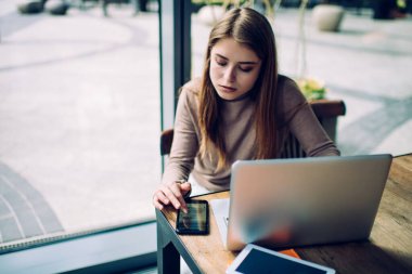 Cafe 'de cam duvarın arkasında dizüstü bilgisayarla otururken cep telefonuna göz atan ciddi genç bir kadının yüksek açısı.