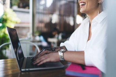 Cropped view of cheerful Caucasian female digital nomad laughing at table on cafe terrace, joyful woman freelancer sitting with laptop computer for doing distance job smiling and rejoicing clipart