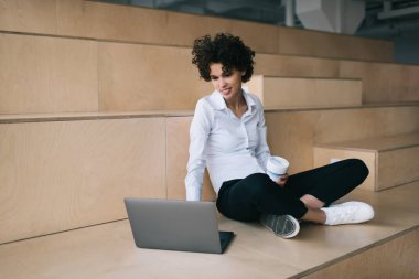 Milenyum Kafkas kızı dizüstü bilgisayardan bir şey izliyor. Modern başarılı kadın kavramı. Genç, gülümseyen kıvırcık kadın. Kahveli. Çalışma odasındaki ahşap bankta oturuyor.