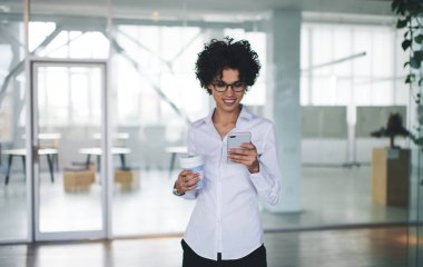 Milenyum Kafkasyalı iş kadını akıllı telefondan izliyor ve kullanıyor. Modern başarılı kadın kavramı. Açık uzay ofisinde elinde kahve fincanıyla gülümseyen kıvırcık kız.