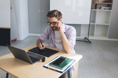 Milenyum Kafkas işadamı laptoptan izliyor ve grafik tabletine bir şeyler yazıyor. Modern başarılı adam kavramı. Gözlüklü genç adam açık uzay ofisinde masada oturuyor.
