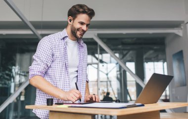 Milenyum Kafkas işadamı laptoptan izliyor ve grafik tabletine bir şeyler yazıyor. Modern başarılı adam kavramı. Geniş ofisteki masasında kablosuz kulaklıklı genç adam.