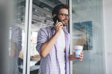 Milenyum Kafkas işadamı elinde kahve fincanıyla akıllı telefondan konuşuyor. Modern başarılı adam kavramı. Genç, gülümseyen, sakallı, gözlüklü ve günlük giysileri olan adam açık uzay ofisinde duruyor.