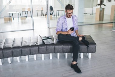 Milenyum Kafkas işadamı akıllı telefonu kullanıyor ve izliyor. Modern başarılı adam kavramı. Deri bankta oturan gözlüklü yakışıklı ve açık uzay ofisinde dizüstü bilgisayarı olan genç adam.