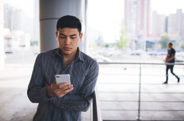 Asyalı genç adam dışarıda akıllı telefondan bir şeyler izliyor. Uzaktan kumanda ve serbest çalışma kavramı. Modern, başarılı bir adamın fikri. Ofis binasının yakınındaki balkonda kulaklıklarda odaklanmış bir erkek.