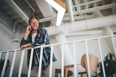 Geniş ofisteki balkonda beyaz adamın akıllı telefondan konuşmasının alt görüntüsü. Uzaktan eğitim ve iş kavramı. Modern, başarılı bir adamın fikri. Gözlüklü genç adam başka tarafa bakıyor.