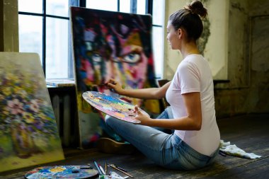 Tavan arası atölyesindeki sanat atölyesinde palet yağı ya da akrilik renkler çizen genç beyaz bir kadın, yerde oturan yetenekli bir kadın ve resim yapmak için hafta sonu boş zaman harcıyor.