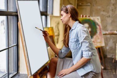 Tablo üzerine soyut resimler çizen sıradan giyinmiş kadın ressam resim sehpasında atölye çalışması yapıyor, yetenekli beyaz ustası hafta sonu sanat stüdyosunda ilham kaynağı yaratıyor.