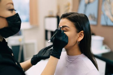 Güzellik uzmanı kuaförde Asyalı bir kızın iğnesiyle kaş mikropateni yapıyor. Kaşlar için yarı kalıcı makyaj. Eldiven ve tıbbi maske takan kadın üstadın kırpılmış görüntüsü. Güzellik konsepti