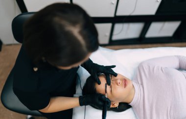 Güzellik uzmanının, güzellik salonunda Asyalı bir kız müşterinin iğnesiyle kaş mikro pateni yaptığı en iyi manzara. Kaşlar için yarı kalıcı makyaj. Kadın üstat eldiven takıyor. Güzellik konsepti
