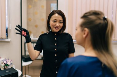 Güzellik uzmanı, modern güzellik salonunda kadın müşteri seçmek için kavanozları rujla gösteriyor. Kalıcı makyaj dudakları prosedürü. Yetişkin Asyalı kadın üstat eldiven giyiyor. Güzellik konsepti