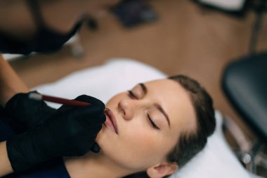 Güzellik uzmanı güzellik salonundaki beyaz kadın müşterinin dudaklarına şekil çiziyor. Kalıcı makyaj dudakları prosedürü. Kadın üstadın eldiven ve tıbbi maske taktığı kısmi görüntü. Güzellik konsepti