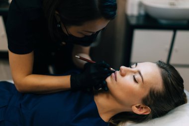 Güzellik uzmanı güzellik salonundaki beyaz kadın müşterinin dudaklarına şekil çiziyor. Kalıcı makyaj dudakları prosedürü. Asyalı kadın üstadın eldiven ve tıbbi maske taktığı görüntüler. Güzellik konsepti