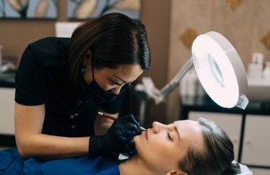 Güzellik uzmanı güzellik salonundaki beyaz kızın dudaklarına şekil çiziyor. Kalıcı makyaj dudakları prosedürü. Eldiven ve tıbbi maske takan Asyalı kadın üstat. Gözleri kapalı bir kadın müşteri. Güzellik konsepti