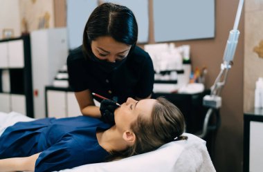 Güzellik uzmanı güzellik salonunda gözleri kapalı beyaz bir kızın dudaklarına sınır çiziyor. Kalıcı makyaj dudakları prosedürü. Eldiven ve tıbbi maske takan Asyalı kadın üstat. Güzellik konsepti