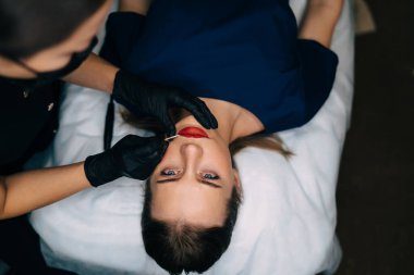 Güzellik uzmanının üst görüşü, Avrupa 'lı kadın müşterinin ahşap çubuğuyla güzellik salonunda kalıcı makyaj dudakları prosedürü uyguluyor. Eldiven ve tıbbi eldiven takan kadın üstadın kırpılmış görüntüsü. Güzellik konsepti