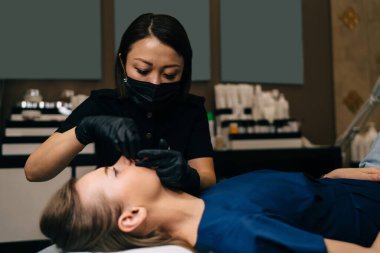 Güzellik uzmanının güzellik salonundaki genç beyaz kız müşterisine kalıcı makyaj dudakları prosedürü uygulaması. Profesyonel Asyalı kadın üstat eldiven ve tıbbi eldiven giyer. Güzellik konsepti