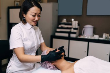 Gülümseyen kadın güzellik uzmanı modern güzellik salonunda kız yüzüne masaj yapıyor. Genç, güzel bir bayan müşteri, gözleri kapalı, tıbbi şapka takarak yatıyor. Vücut ve cilt bakımı kavramı