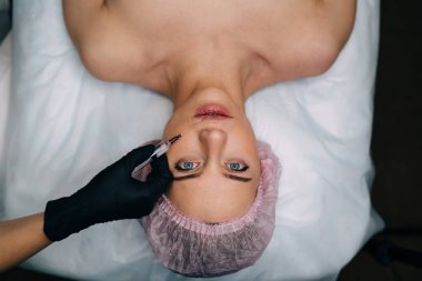 Güzellik uzmanının güzellik salonundaki kıza şırıngayla botoks yaparken çekilmiş görüntüsü. Sağlık şapkası takmış kameraya bakan beyaz kadın müşterinin en iyi görüntüsü. Güzellik konsepti. Yüz gerdirme