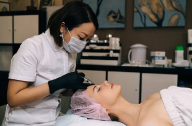 Asyalı kadın güzellik uzmanı güzellik salonundaki kıza şırınga ile botoks yapıyor. Tıbbi başlık takan, gözleri kapalı, beyaz, genç, çekici bir bayan müşteri. Güzellik konsepti. Yüz gerdirme