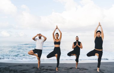 Spor elbiseli genç bir adam ve kadın yoga pozisyonunda duruyor. Açık havada nefes alıyor ve eğleniyor. Bir grup Yogi, Asana Sahili 'ndeki sahil eğitiminde ilham ve dengeyi hissediyor.