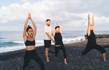 Sahil şeridinde birlikte denge uygulaması yaparken günlük spor kıyafetlerinde asana erkek ve dişilerine eğitim veren Kafkas yogası. Deniz kıyısındaki danışman eğitmenlerin tavsiyeleri.