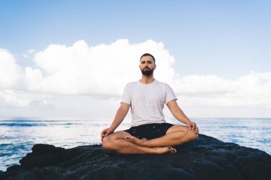Sportif giyinen sakin erkek portresi lotus pozisyonunda oturur ve okyanus kenarında sabah meditasyonu sırasında kameraya bakar. Genç beyaz adam, doğanın kıyı şeridinde yoga yaparken dinlenir.