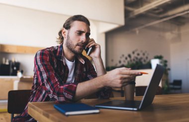 Milenyum Kafkasyalı akıllı telefonla konuşuyor ve iş sırasında dizüstü bilgisayardan izliyor. Modern başarılı adam kavramı. Genç odaklı erkek girişimci açık uzay ofisindeki ahşap masada oturuyor.