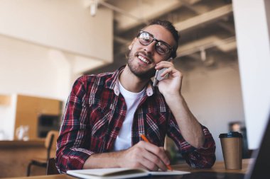 Milenyum Kafkasyalı akıllı telefonla konuşuyor ve iş sırasında deftere yazı yazıyor. Modern başarılı adam kavramı. Gözlüklü genç adam kameraya bak ve açık uzay ofisindeki masaya otur.