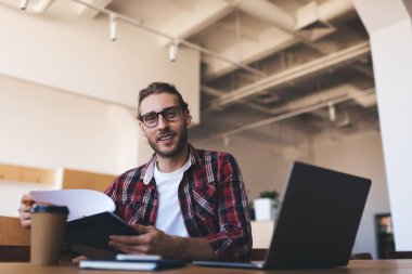 Milenyum Kafkasyalı iş sırasında kameraya bakıyor. Modern başarılı adam kavramı. Gözlüklü genç girişimci açık uzay ofisindeki ahşap masada oturuyor.