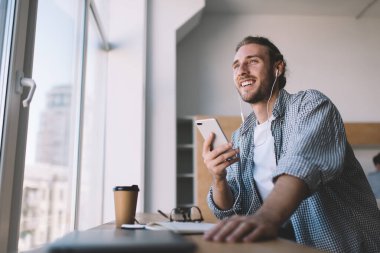 Milenyum Kafkasyalı iş adamı akıllı telefonuyla kulaklıkla müzik dinliyor. Modern başarılı adam kavramı. Genç memnun erkek girişimci açık uzay ofisindeki pencere eşiğine bakıyor.