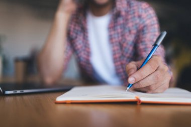 Bulanık bir adamın not defterine kalemle bir şeyler yazdığı seçmeli bir odak noktası. Modern başarılı adam kavramı. Erkek girişimci, ofisinde bilgisayarını açıp ahşap masada çalışıyor.