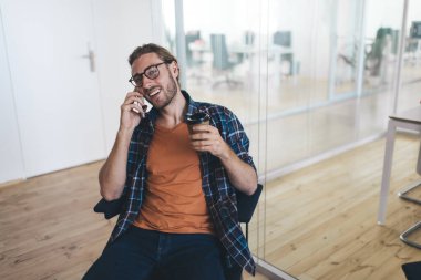 Milenyum Kafkasyalı işadamı elinde kahveyle akıllı telefondan konuşuyor. Modern başarılı adam kavramı. Gözlüklü genç bir girişimci açık uzay ofisindeki kameraya bakıyor.