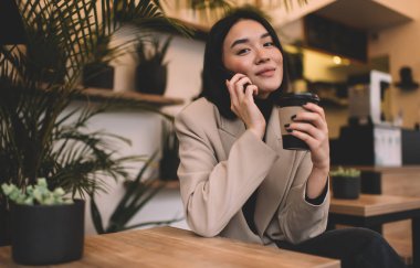 Milenyum Asyalı çekici bir kız akıllı telefondan konuşuyor ve kafede kahve içiyor. Genç esmer kadın masada oturuyor ve kameraya bakıyor. Dinlenme, boş zaman ve boş zaman. Modern kadın yaşam tarzı